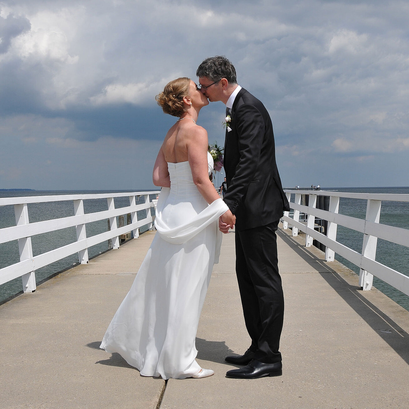 Hochzeitspaar auf der Seebrücke vor dem Maritim Hotel Timmendorfer Strand, das sich bei bewölktem Himmel einen Kuss gibt.