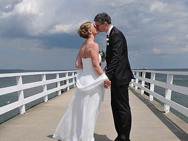 Hochzeitspaar auf der Seebrücke vor dem Maritim Hotel Timmendorfer Strand, das sich bei bewölktem Himmel einen Kuss gibt.