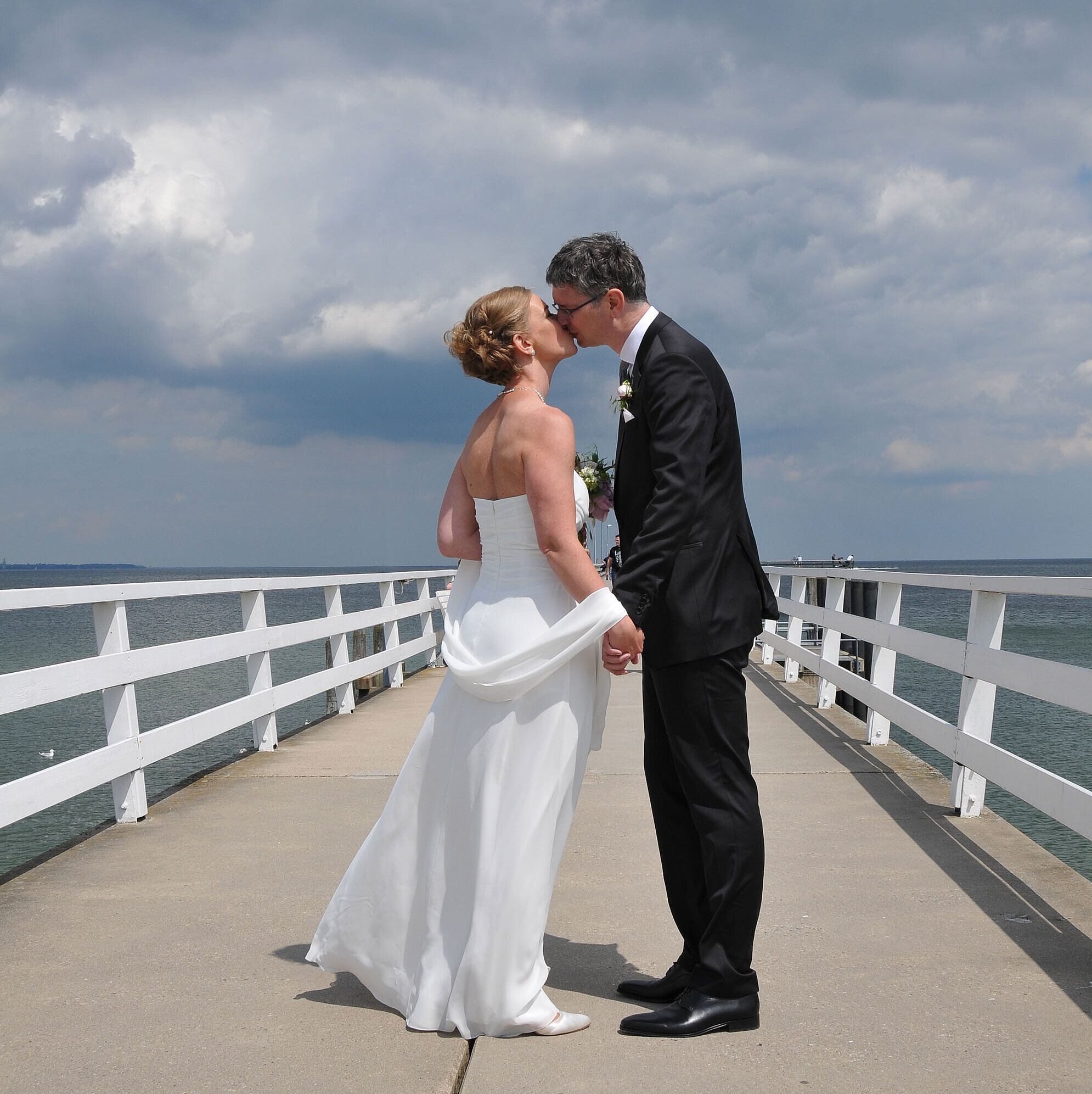 Hochzeit am Meer Hochzeitspaar auf der Seebrücke vor dem Maritim Hotel Timmendorfer Strand, das sich bei bewölktem Himmel einen Kuss gibt.
