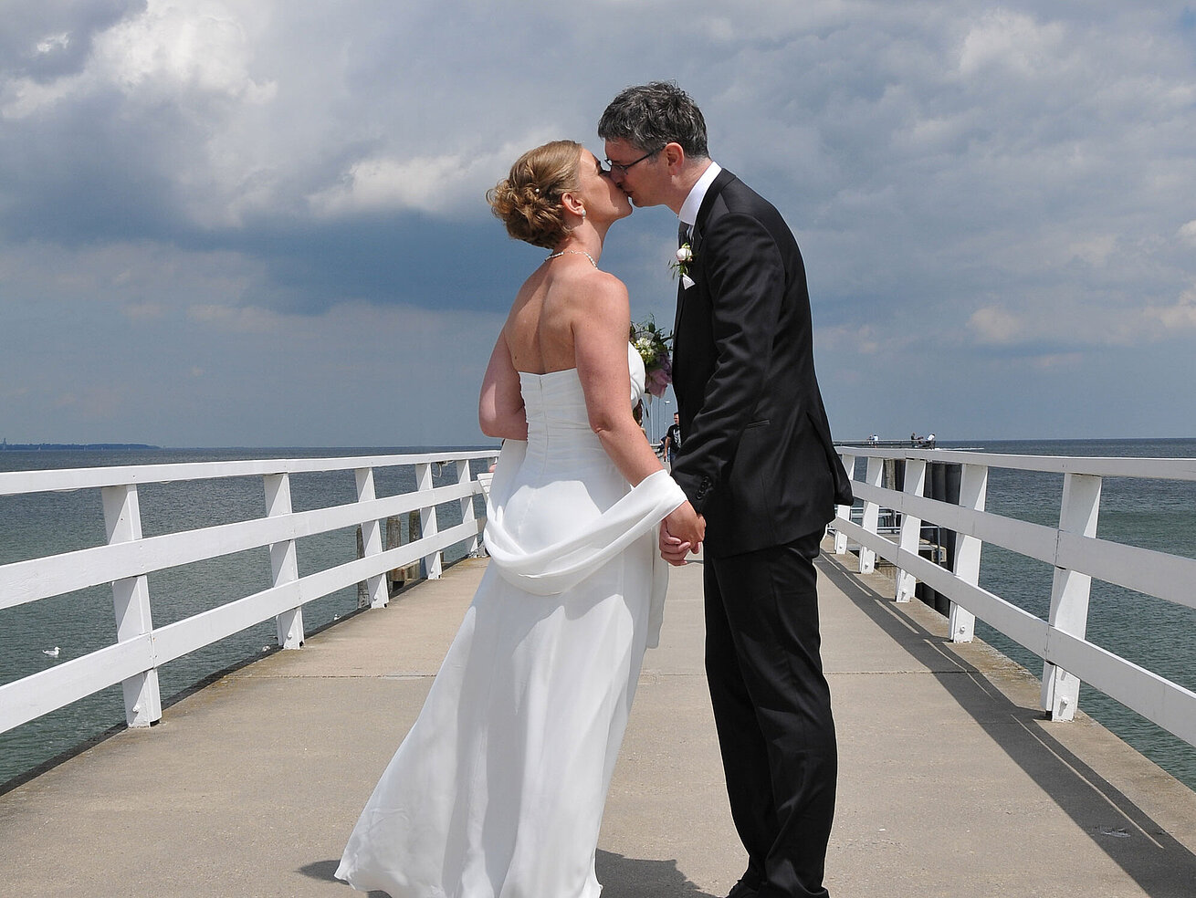 Hochzeitspaar auf der Seebrücke vor dem Maritim Hotel Timmendorfer Strand, das sich bei bewölktem Himmel einen Kuss gibt.