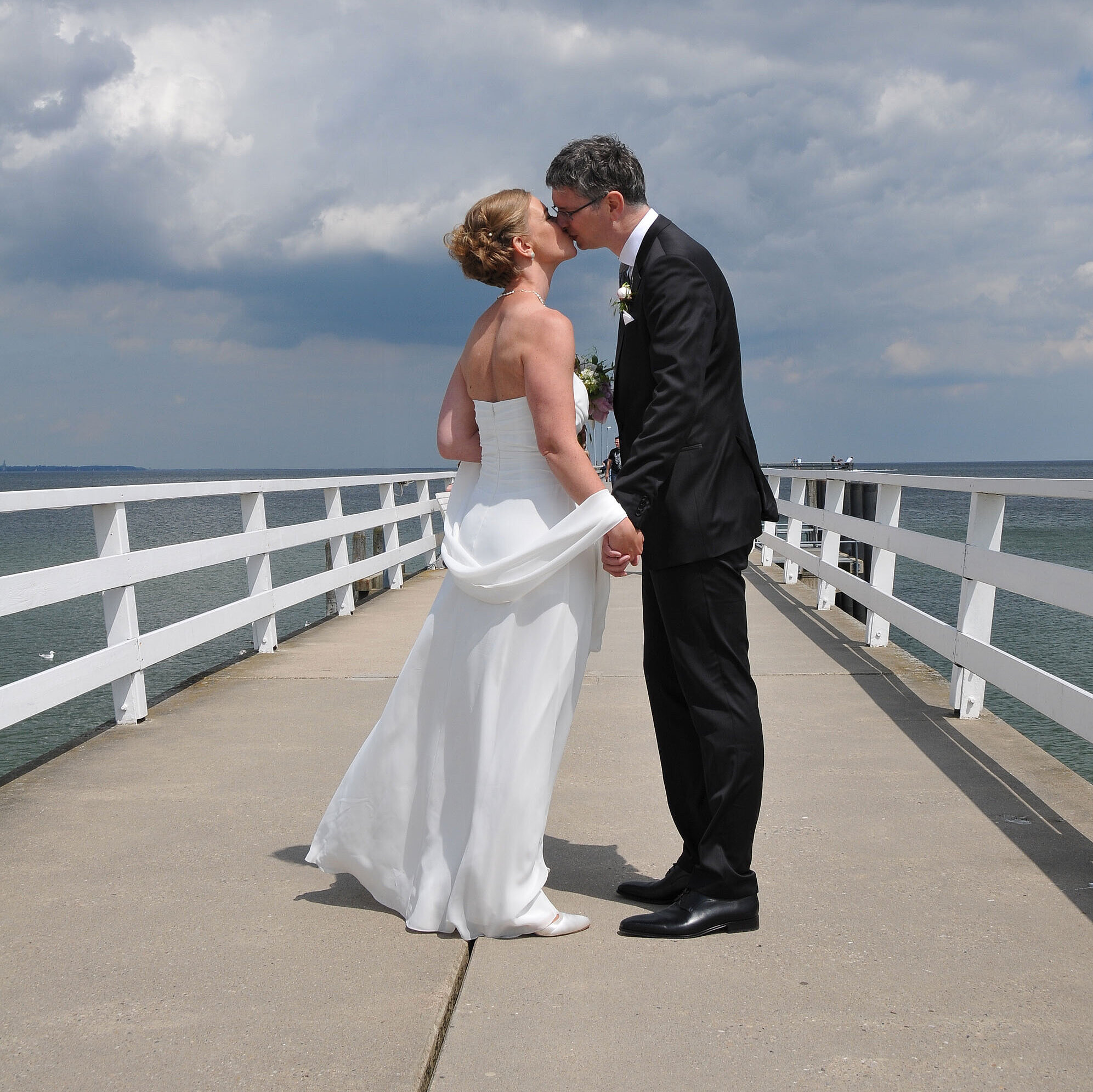Hochzeitspaar auf der Seebrücke vor dem Maritim Hotel Timmendorfer Strand, das sich bei bewölktem Himmel einen Kuss gibt.