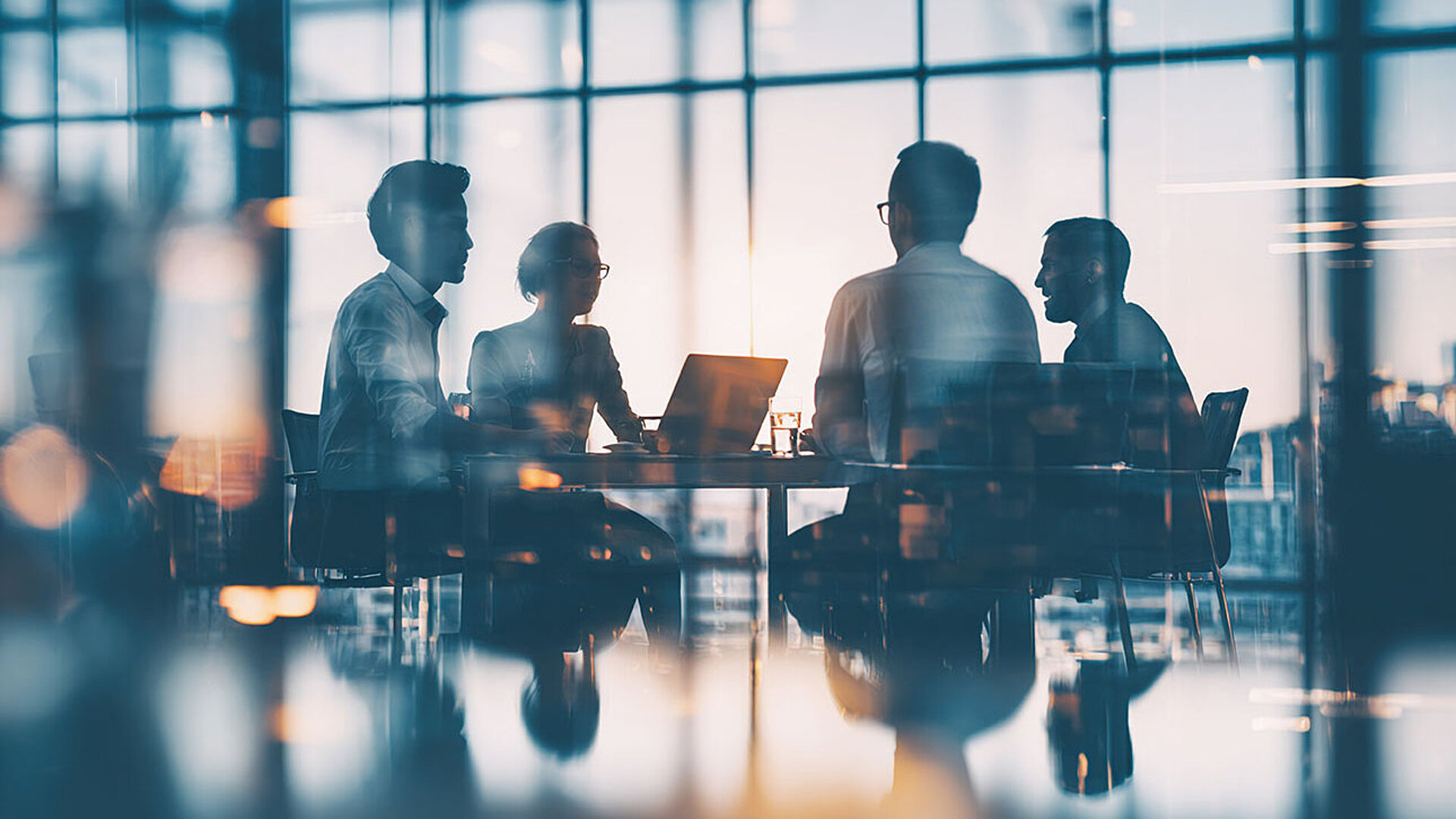 Vier Personen sitzen in einem modernen Konferenzraum mit großen Fenstern und besprechen sich am Tisch.