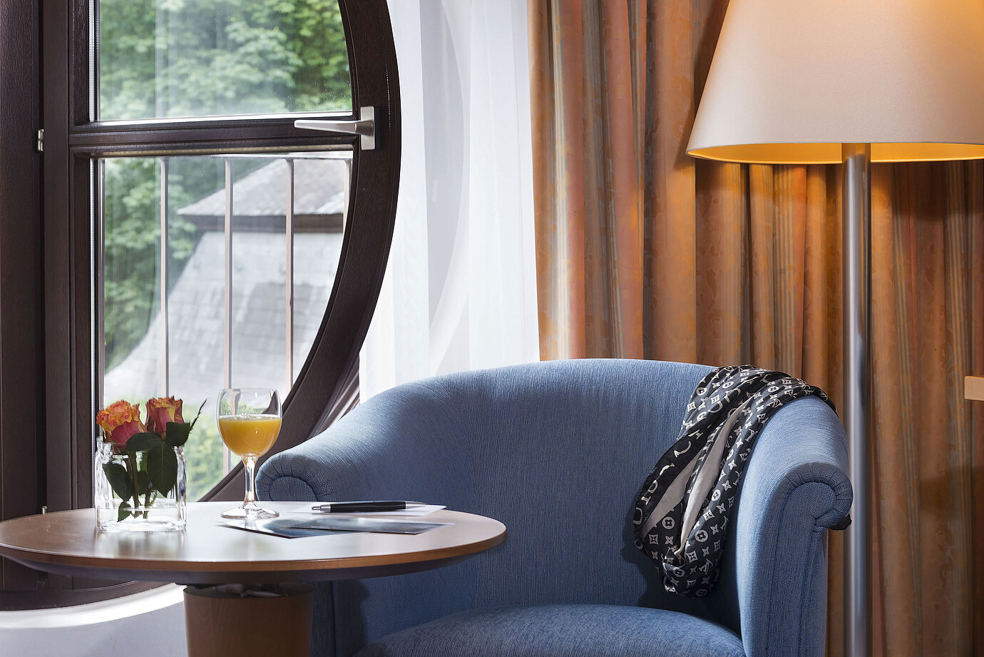  Gemütlicher Sitzbereich im Classic Zimmer des Maritim Hotel Fulda mit blauem Sessel, rundem Fenster, Blumen und Orangensaft.
