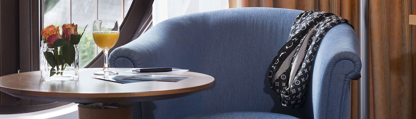  Gemütlicher Sitzbereich im Classic Zimmer des Maritim Hotel Fulda mit blauem Sessel, rundem Fenster, Blumen und Orangensaft.