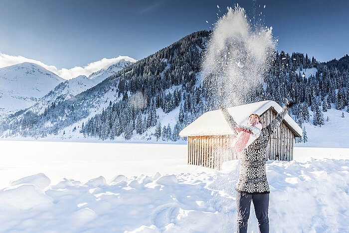 Person wirft Schnee in die Luft vor verschneiten Bergen und einer Hütte in strahlender Winterlandschaft.