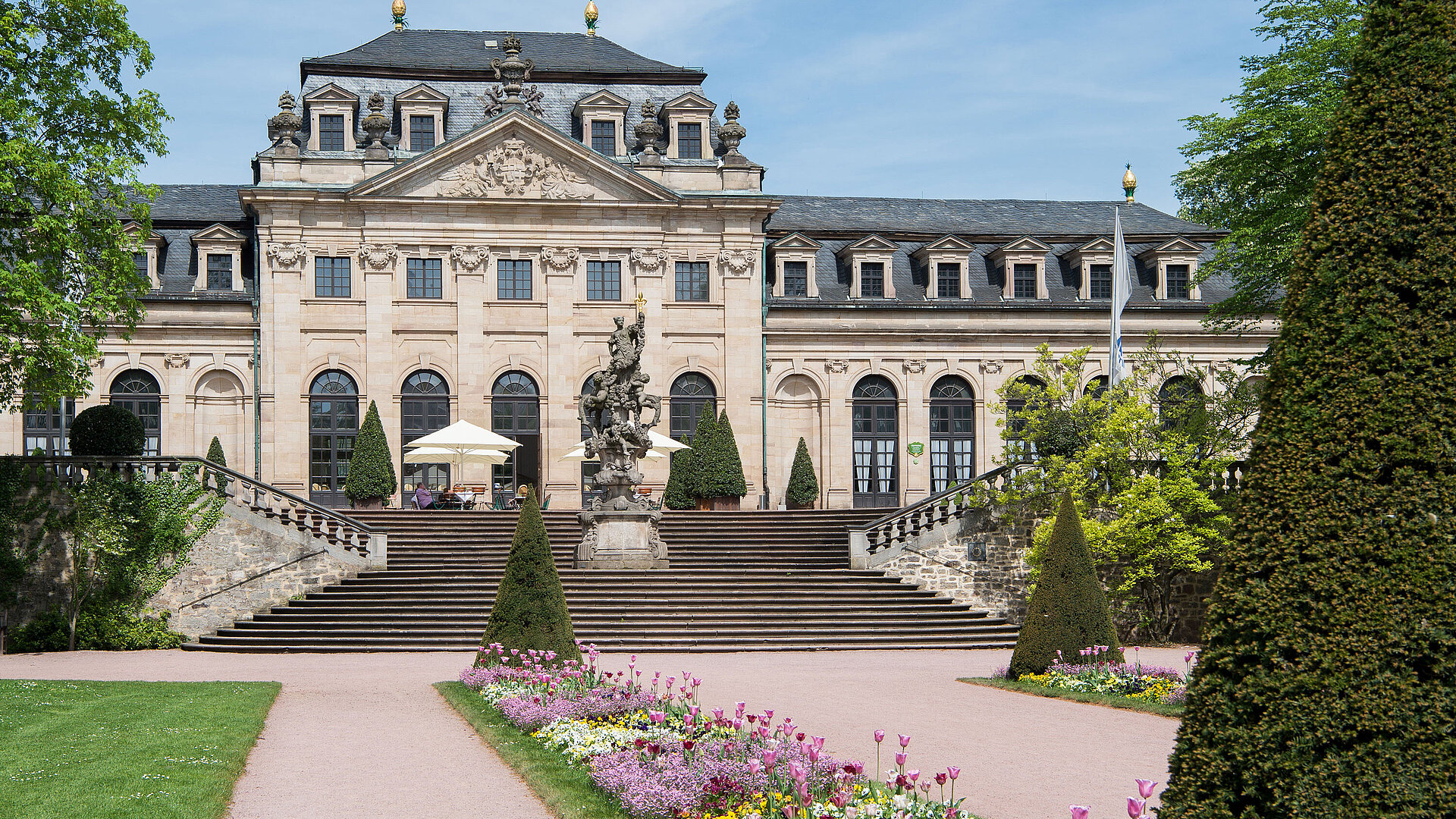 Die historische Orangerie in Fulda mit barocker Fassade, Treppe und blühendem Garten im Vordergrund.