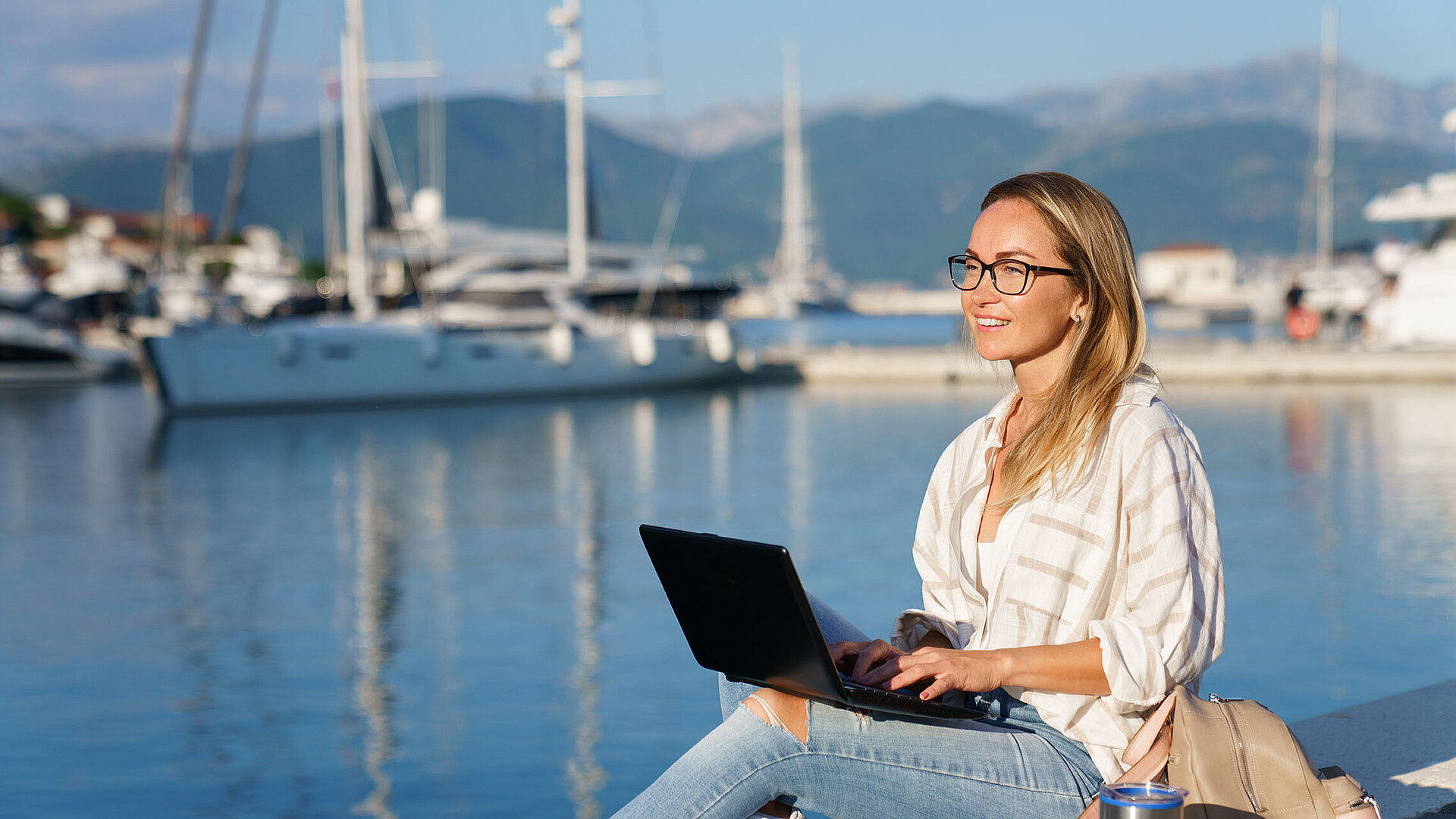 Frau arbeitet mit Laptop am Hafen vor Segelbooten und ruhigem Wasser bei Sonnenschein.