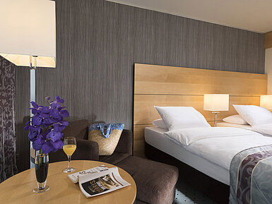 Classic Zimmer im Maritim Hotel Düsseldorf mit elegantem Doppelbett, gemütlicher Sitzecke, frischer Blume und Lesematerial.