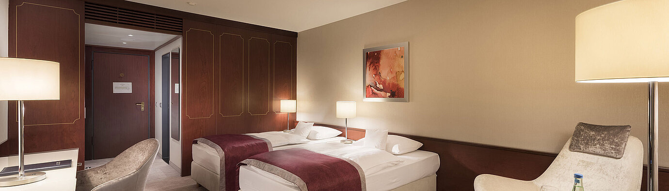Comfort Zimmer im Maritim Hotel Bonn, ausgestattet mit zwei Einzelbetten, Sessel, Schreibtisch und moderner Beleuchtung.