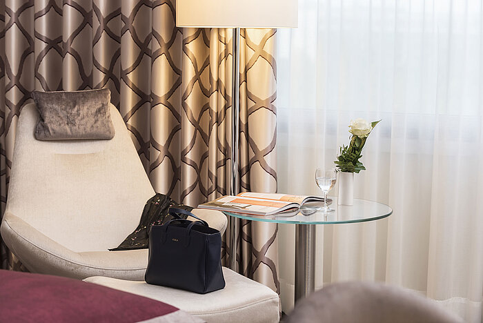 Detail eines Classic Zimmers im Maritim Hotel Bonn mit gemütlichem Sessel, Vorhang, Tisch, Glas, Rose und einer Tasche.