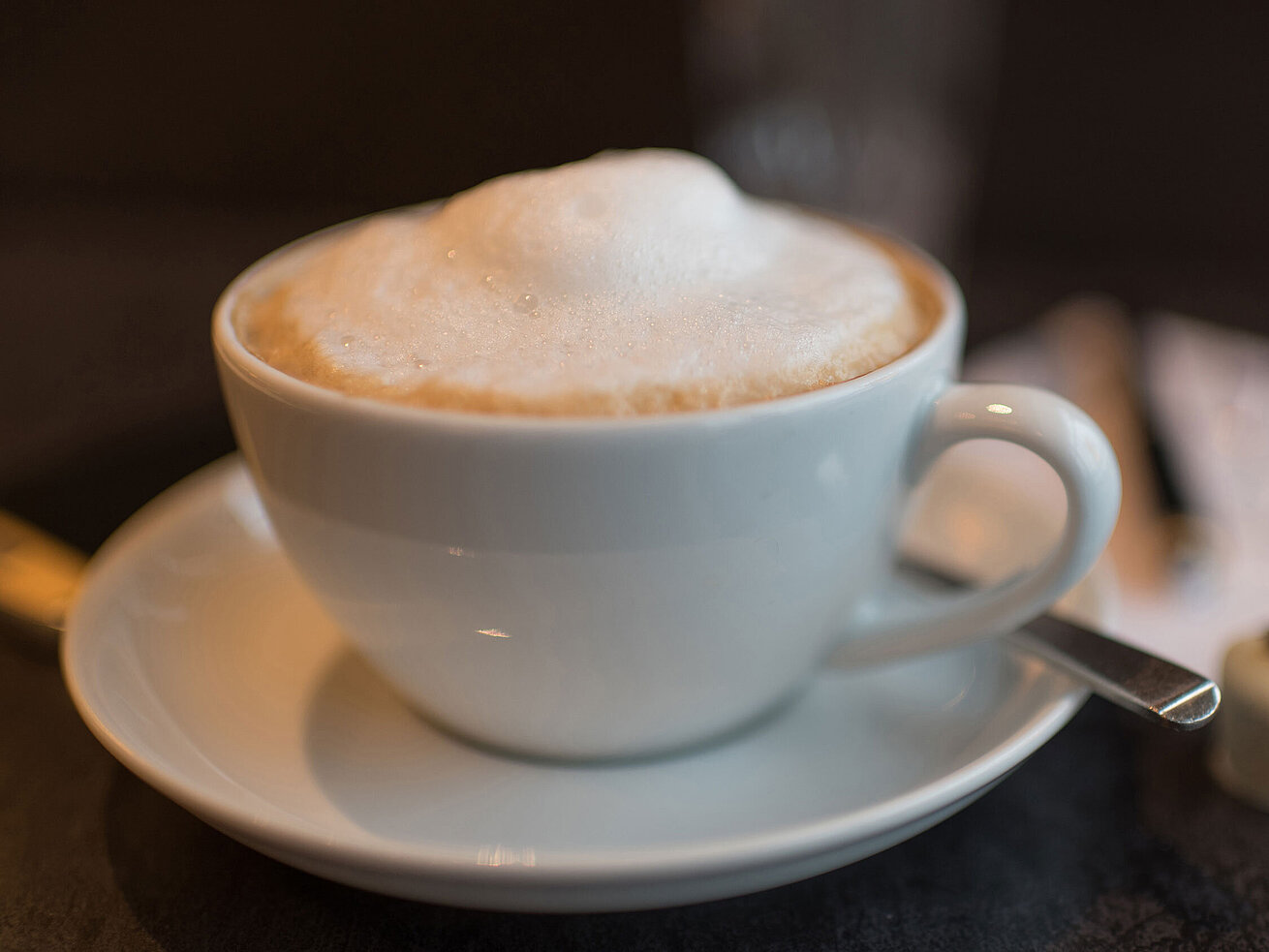 Tasse Cappuccino mit cremigem Milchschaum auf Untertasse, daneben Pralinen auf dunkler Tischplatte.