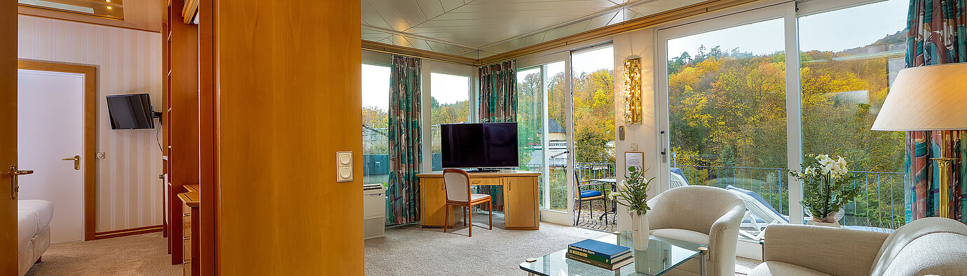 Wintergarten Suite Helles Hotelsuite-Wohnzimmer mit Sesseln, Glastisch, TV und großer Fensterfront mit Blick auf die Umgebung.