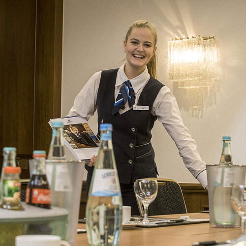 Freundliche Servicemitarbeiterin im Salon Lenné des Maritim Hotels Bonn mit Getränken und Tagungsunterlagen auf dem Tisch.