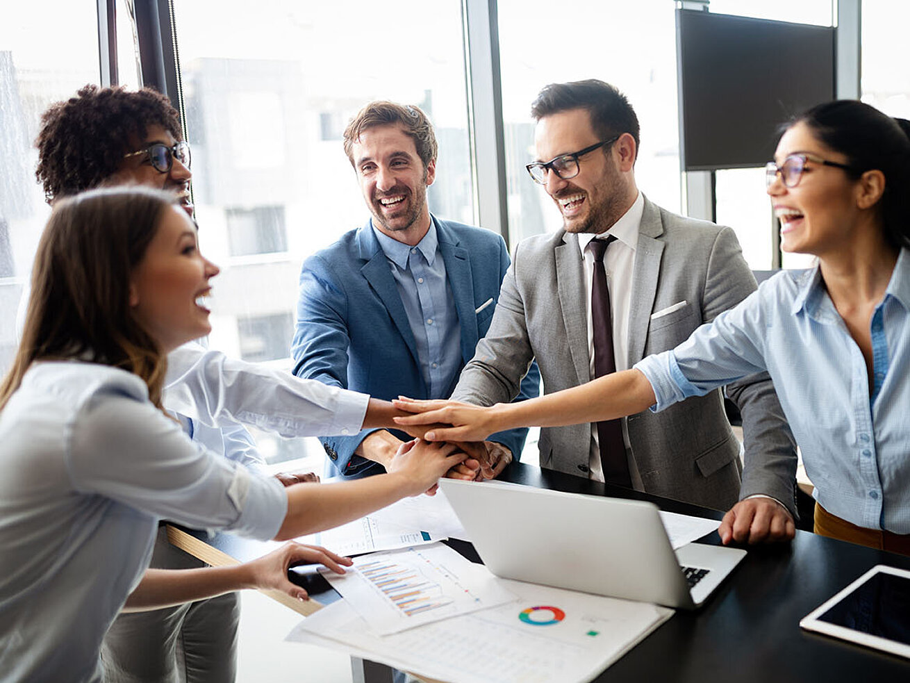 © NDABCREATIVITY - AdobeStock.com Motiviertes Business-Team im Büro legt Hände zusammen, Meeting mit Laptop und Unterlagen am Konferenztisch