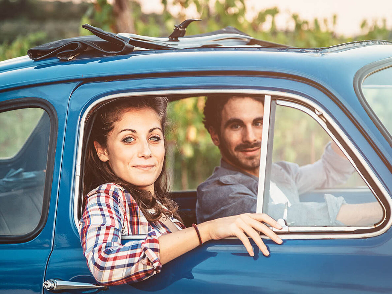 © loreanto - AdobeStock.com Junges Paar sitzt in einem blauen Oldtimer-Auto und blickt lächelnd aus dem Fenster