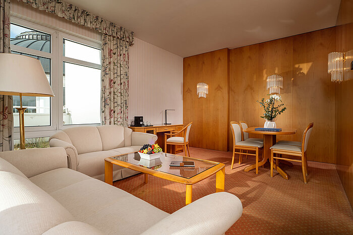 Junior Suite Junior Suite mit hellem Wohnbereich, Sofa, Glastisch, Essplatz und gemütlicher Einrichtung im Maritim Hotel.