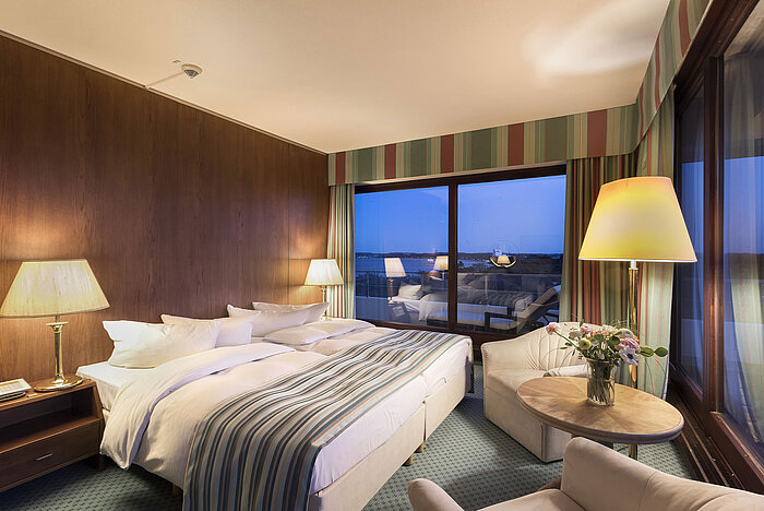 Superior Zimmer im Maritim Seehotel Timmendorfer Strand mit Doppelbett, Sitzbereich, Stehlampen und nächtlichem Balkonblick.