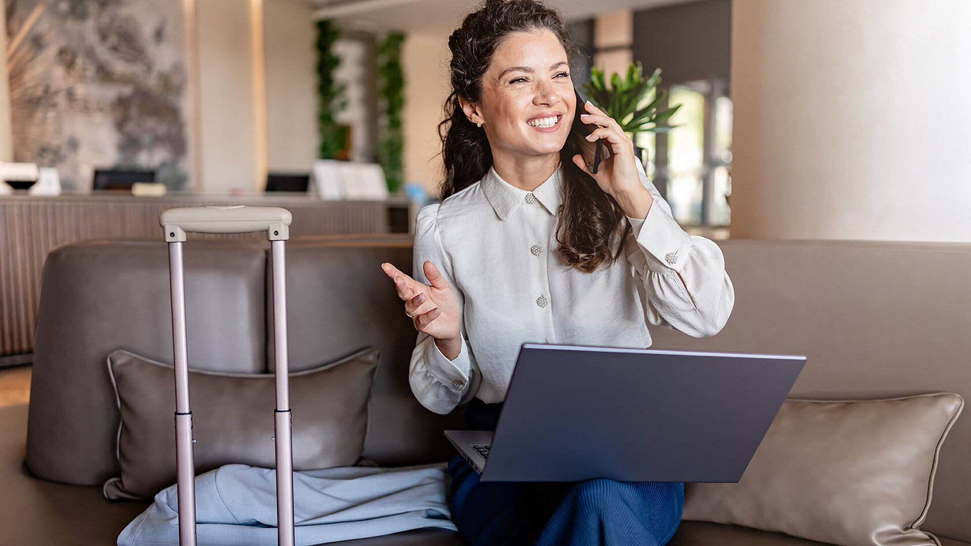 Geschäftsfrau sitzt mit Laptop auf dem Sofa in einer Hotel-Lobby, telefoniert lächelnd und hat einen Koffer neben sich