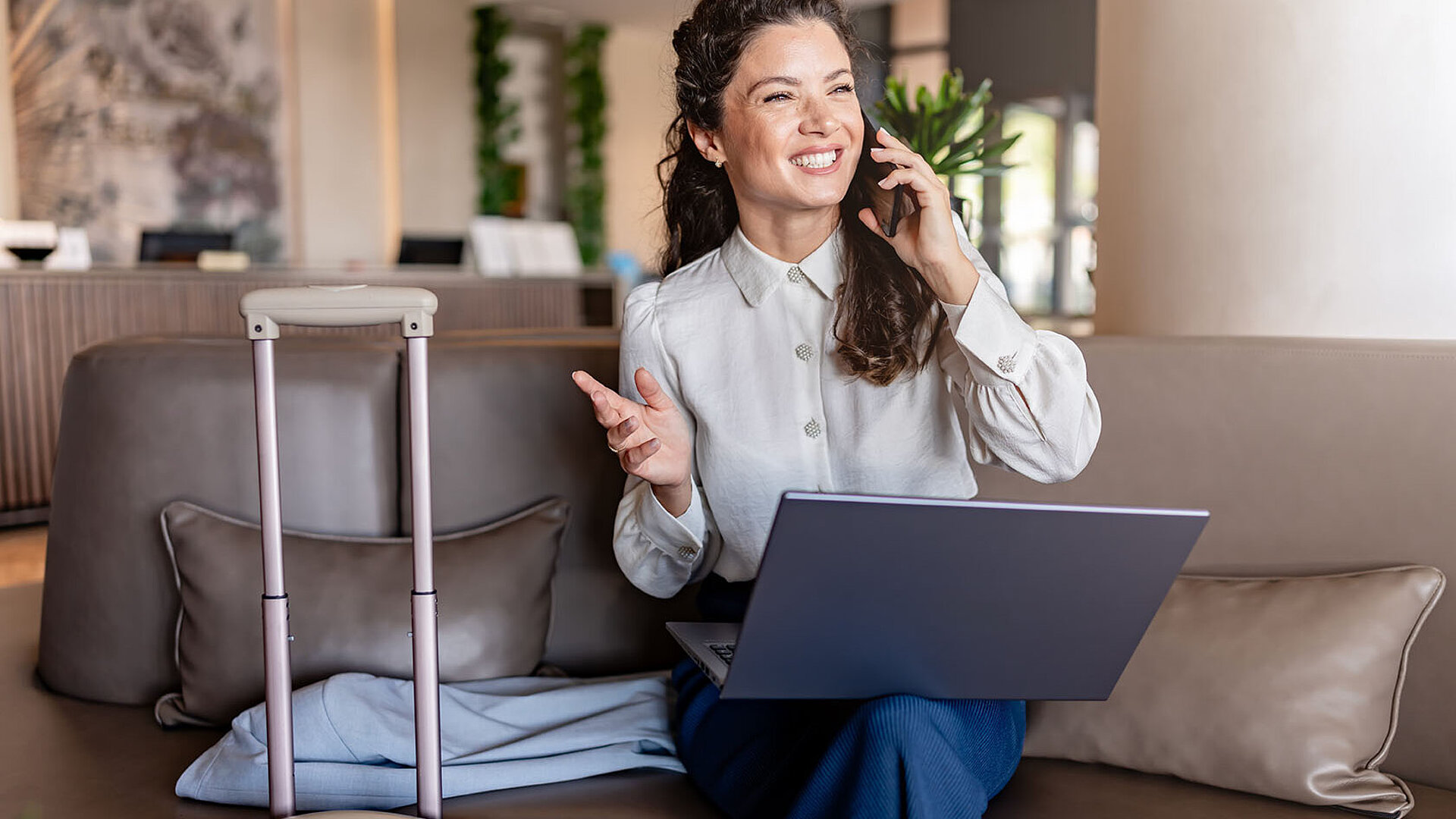 Geschäftsfrau sitzt mit Laptop auf dem Sofa in einer Hotel-Lobby, telefoniert lächelnd und hat einen Koffer neben sich