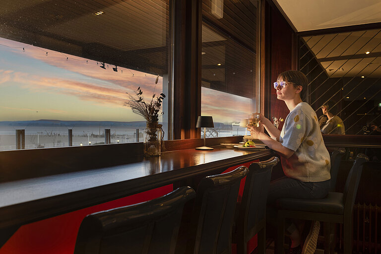 Gast sitzt mit Getränk am Fenster und blickt auf den Sonnenuntergang im Maritim Hotel Travemünde