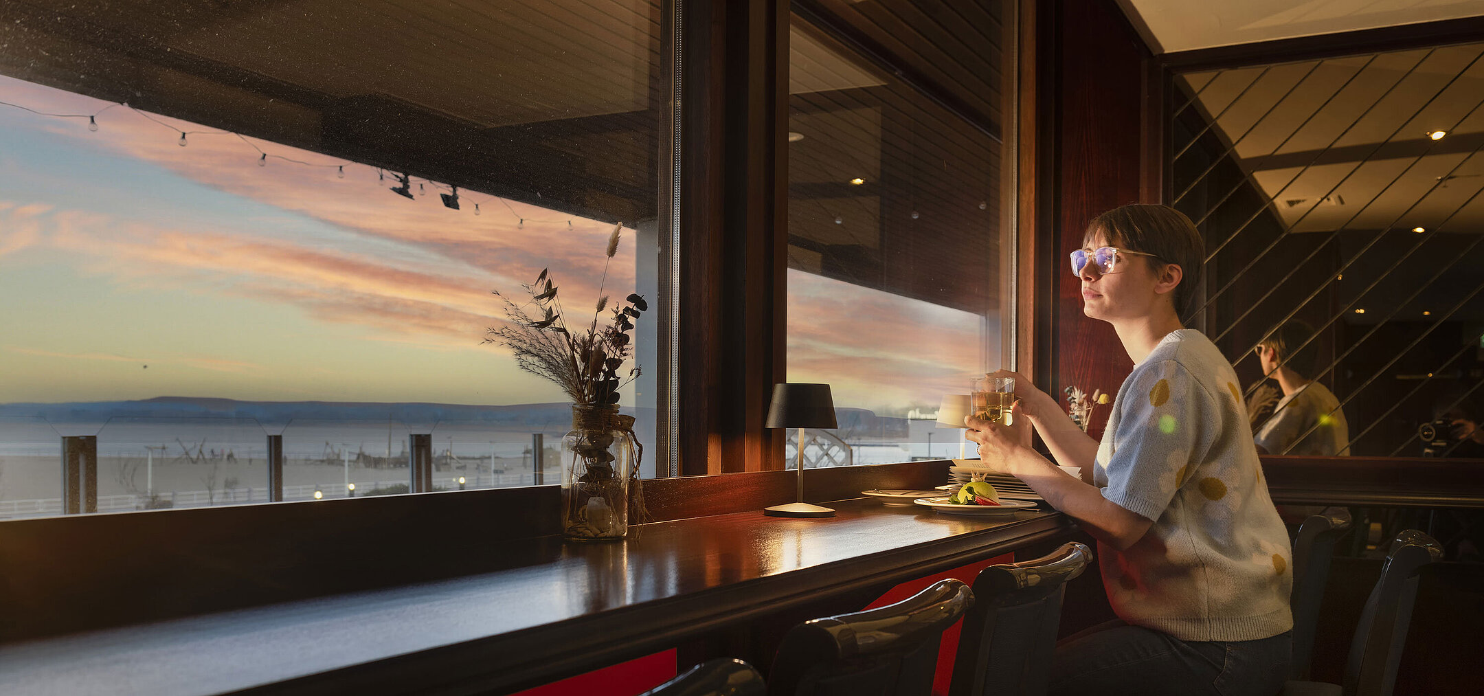 Gast sitzt mit Getränk am Fenster und blickt auf den Sonnenuntergang im Maritim Hotel Travemünde