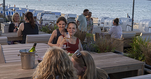  Gäste genießen Wein in der Strandbar des Maritim Seehotel Timmendorfer Strand mit Blick auf Strandkörbe und das Meer.