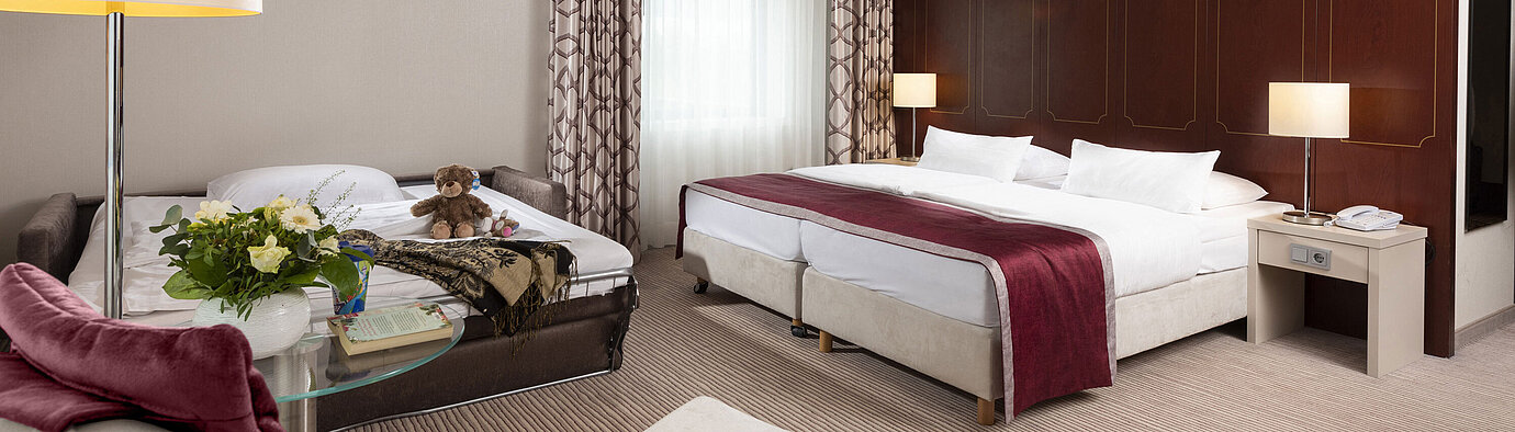 Comfort Familienzimmer im Maritim Hotel Bonn mit großem Doppelbett, ausziehbarem Sofa, Sessel und Blumenarrangement.