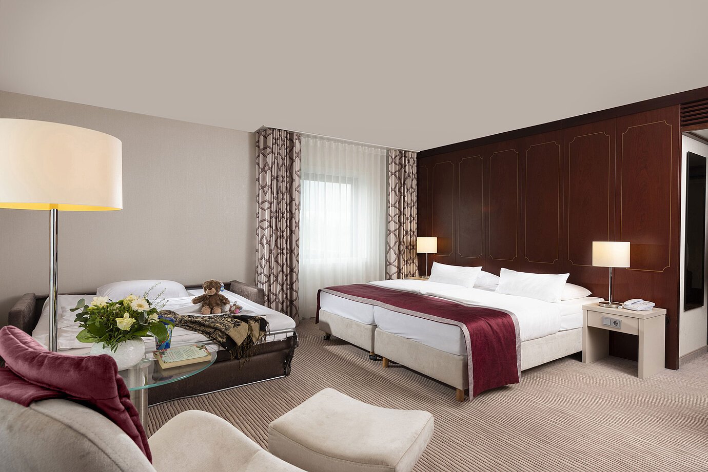 Comfort Familienzimmer im Maritim Hotel Bonn mit großem Doppelbett, ausziehbarem Sofa, Sessel und Blumenarrangement.