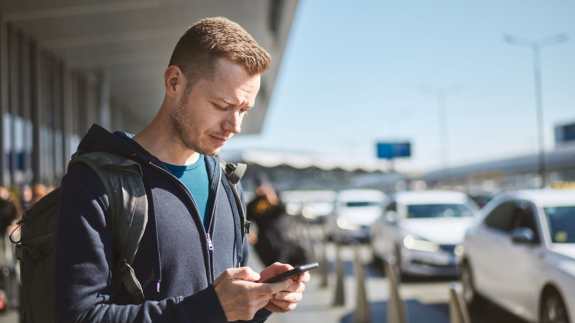 Mann mit Rucksack nutzt Smartphone vor Flughafen, bereit für Transfer oder Taxi