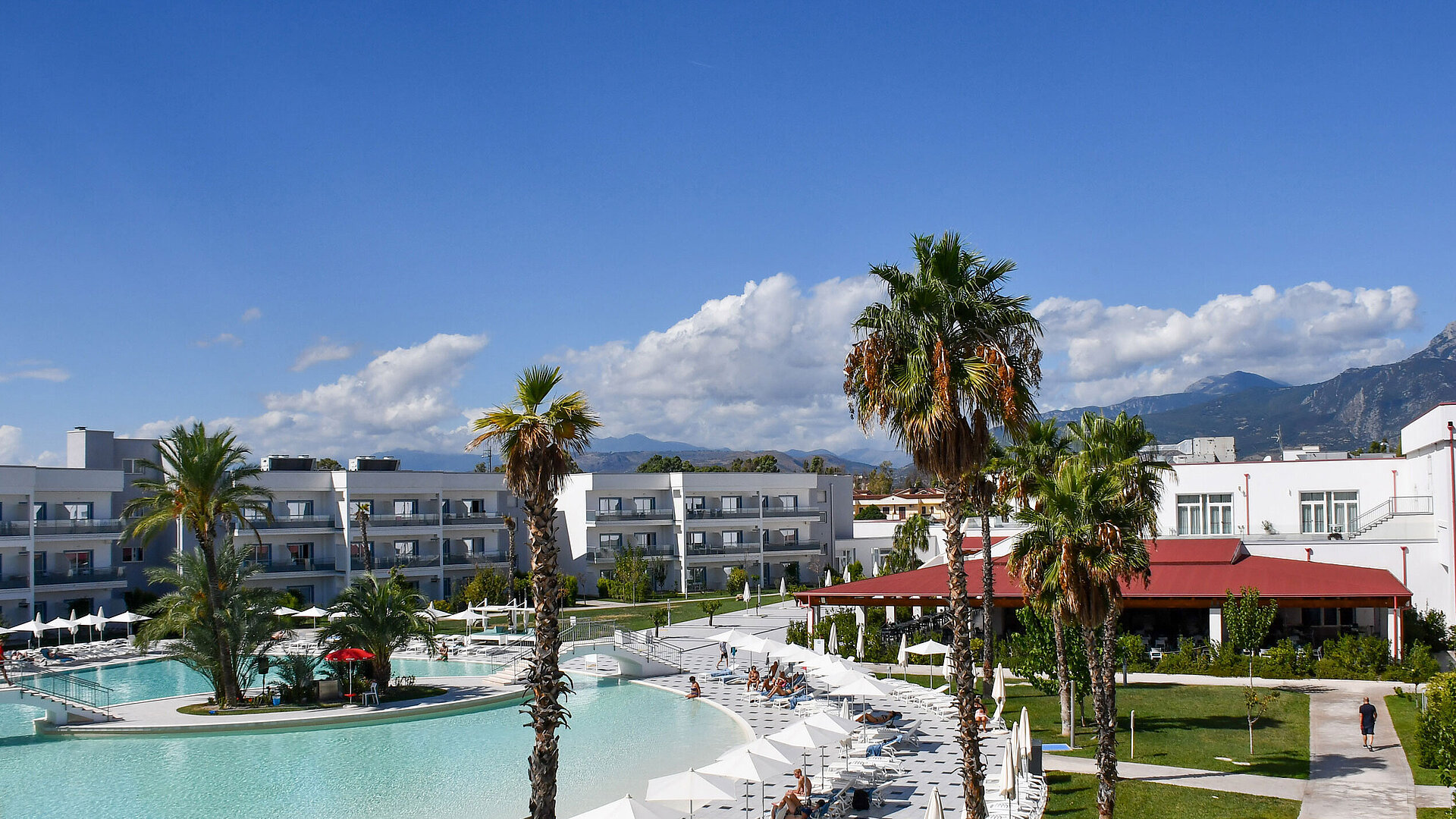 Panorama des Maritim Resort Calabria mit Pool, Palmen, weißen Gebäuden und Bergkulisse im Hintergrund bei klarem Himmel.