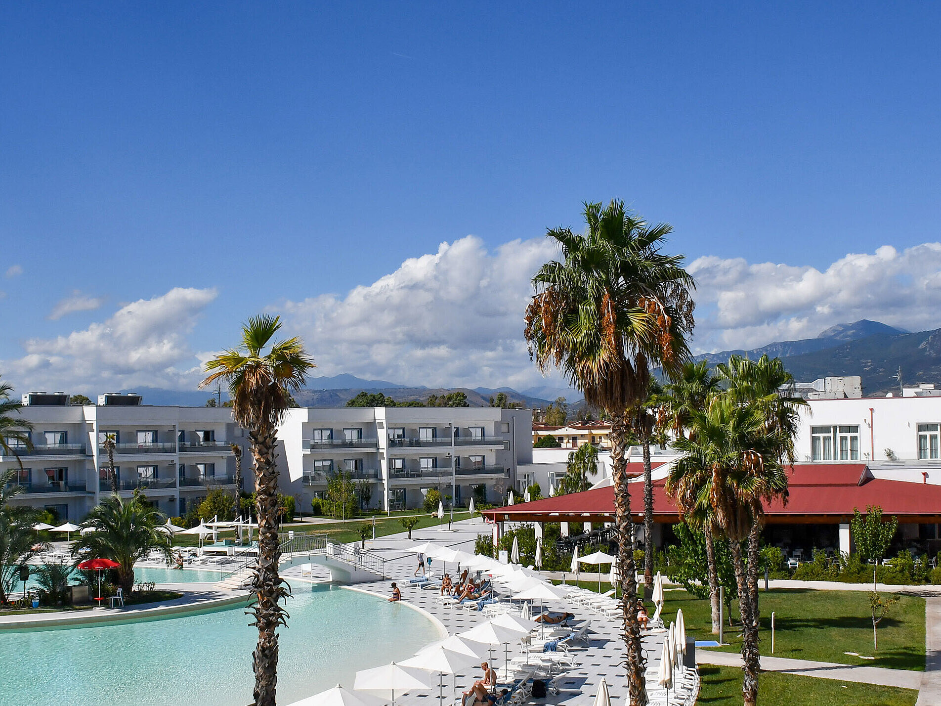 Panorama des Maritim Resort Calabria mit Pool, Palmen, weißen Gebäuden und Bergkulisse im Hintergrund bei klarem Himmel.