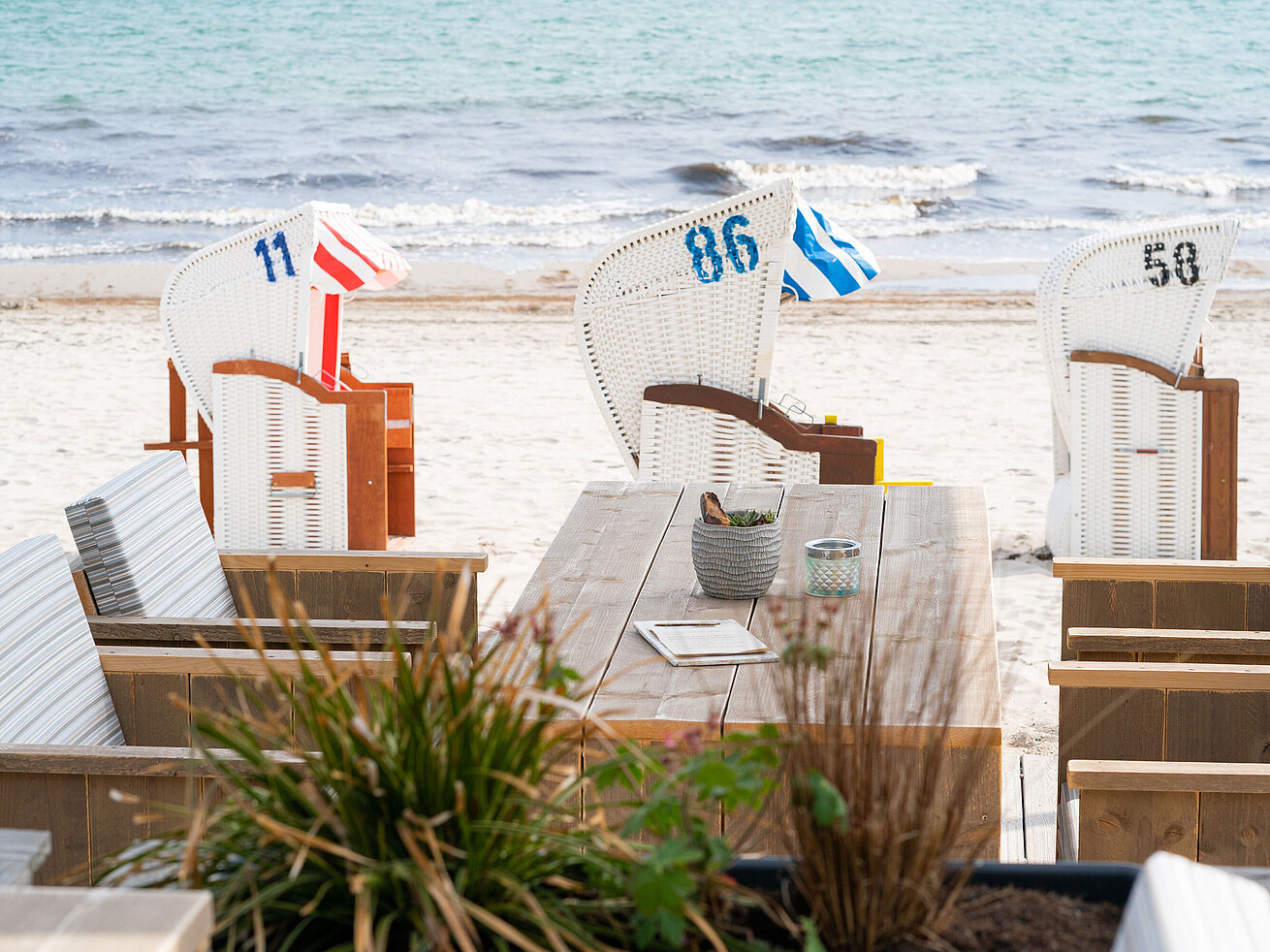 Strandkörbe am weißen Sandstrand, mit Blick auf die ruhige Ostsee und einem gemütlichen Holztisch in der Beach Lounge.