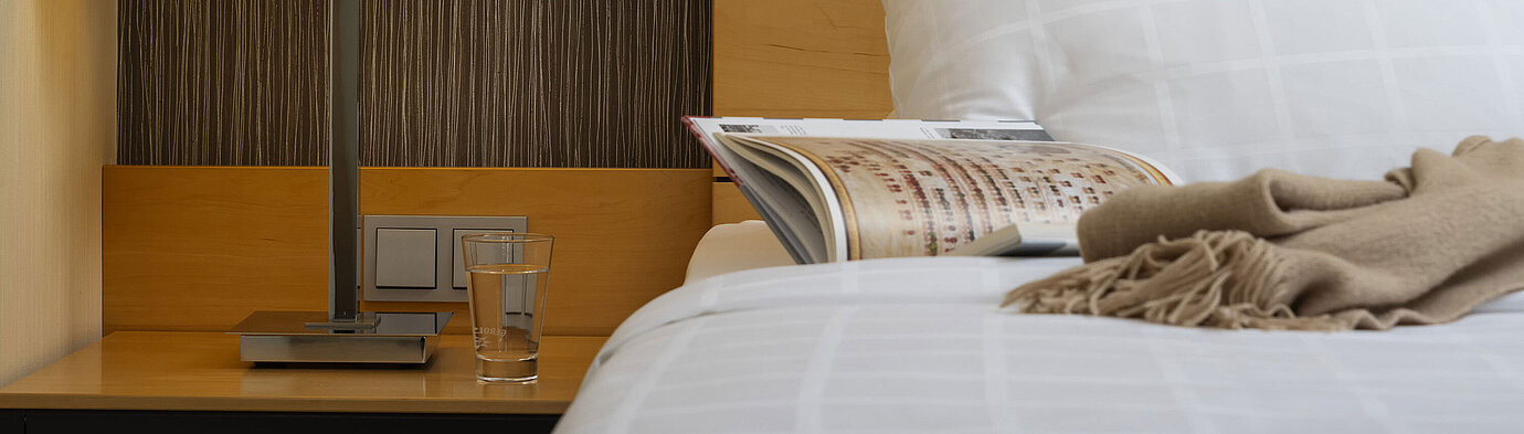 Detailaufnahme eines Nachttisches im Maritim Hotel Düsseldorf mit Lampe, Glas Wasser und Buch auf einem Bett.
