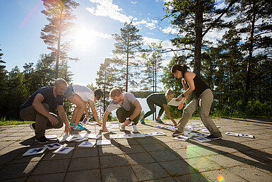 © Tyler Olson AdobeStock.com Gruppe von Erwachsenen beim Outdoor-Teambuilding macht gemeinsame Fitnessübung im Wald bei Sonnenschein