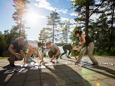 Gruppe von Erwachsenen beim Outdoor-Teambuilding macht gemeinsame Fitnessübung im Wald bei Sonnenschein