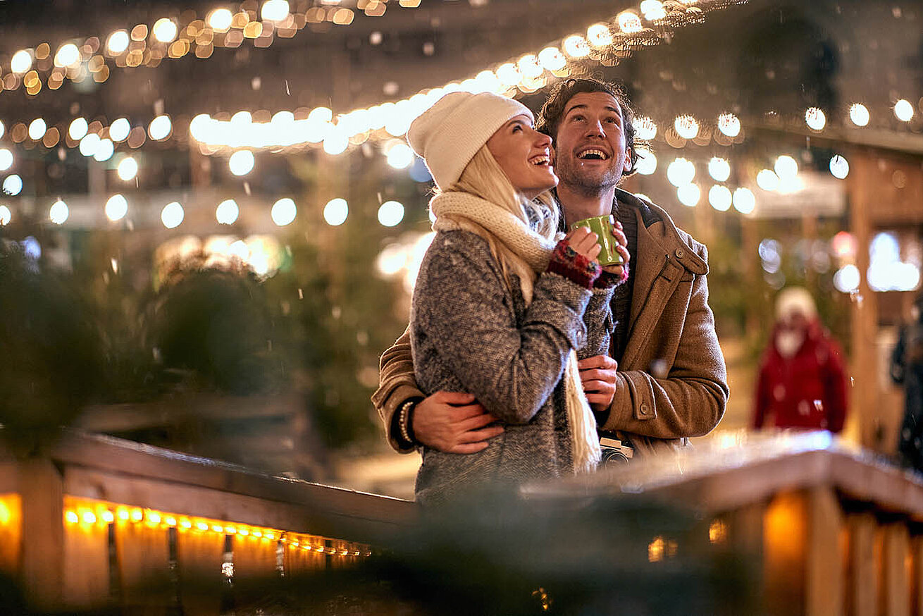 Paar genießt einen winterlichen Abend auf dem Weihnachtsmarkt, umgeben von warmen Lichtern und festlicher Stimmung.