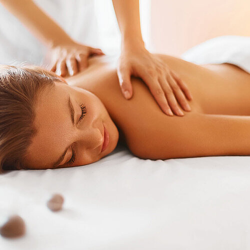 © puhhha - AdobeStock.com Frau genießt entspannende Rückenmassage im Spa mit warmem Licht und Steinen