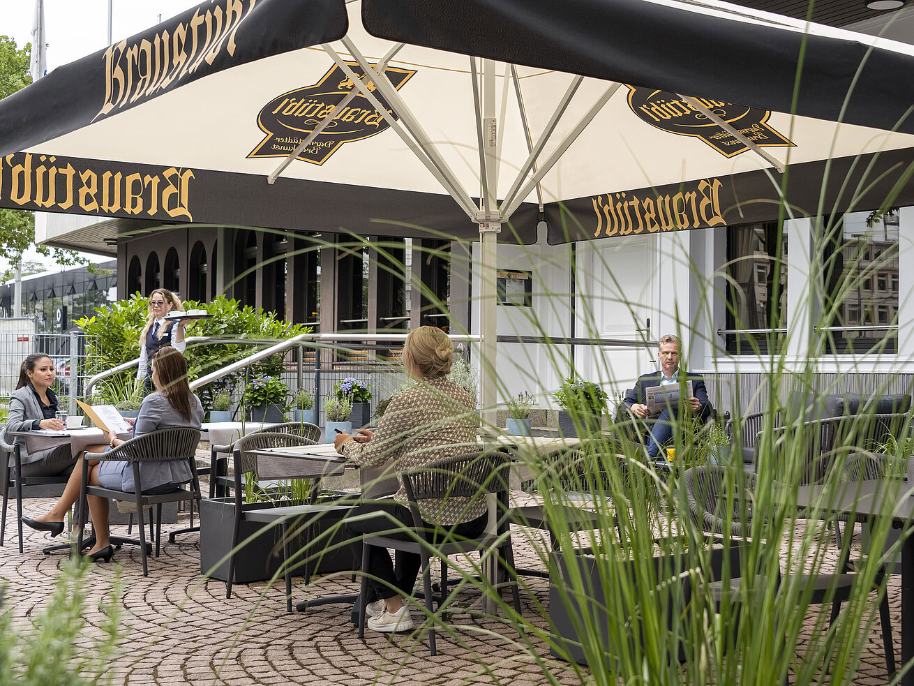 Terrasse Gäste genießen eine Mahlzeit auf der Terrasse des Maritim Hotels Darmstadt unter großen Sonnenschirmen mit Brauhaus-Logo.