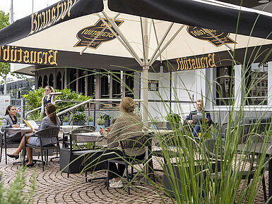 Gäste genießen eine Mahlzeit auf der Terrasse des Maritim Hotels Darmstadt unter großen Sonnenschirmen mit Brauhaus-Logo.