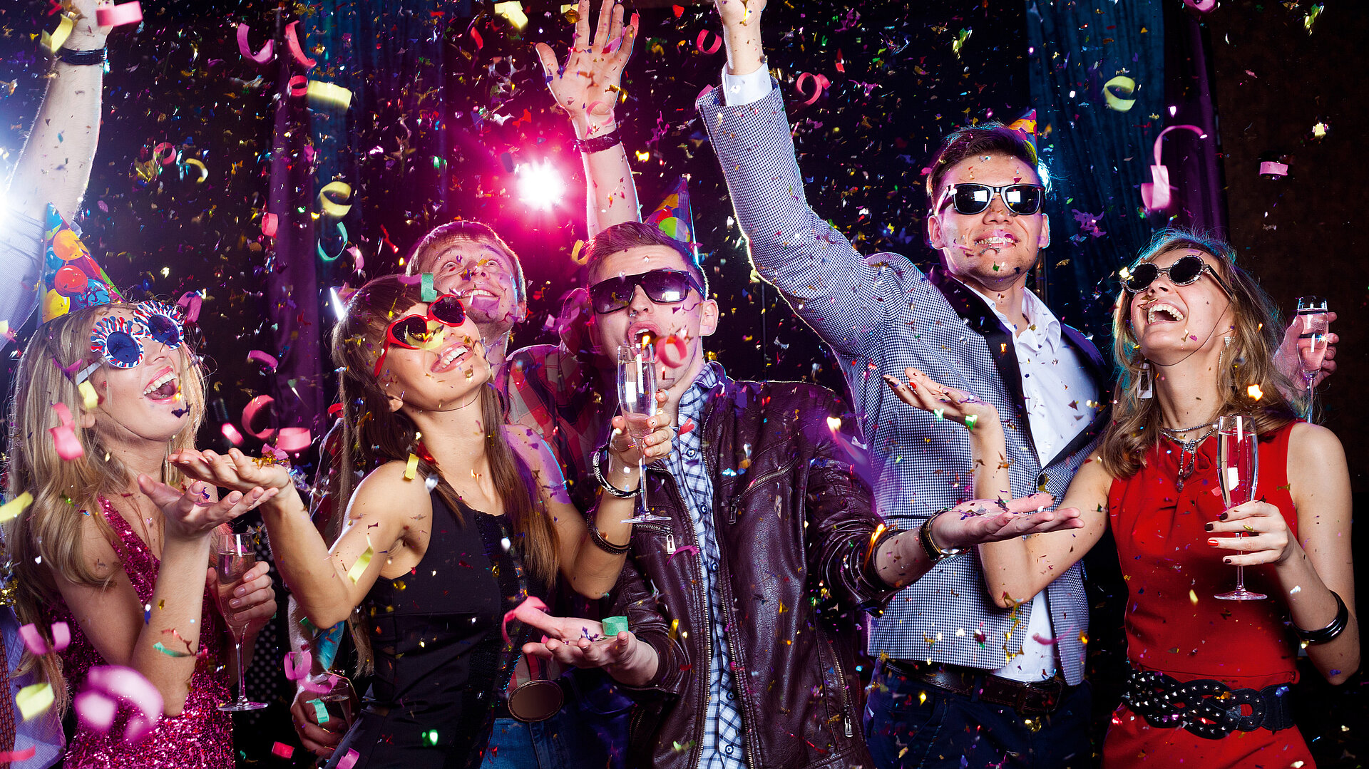 © yanlev - AdobeStock.com Fröhliche junge Leute feiern mit Partyhüten, Sonnenbrillen, Sektgläsern und Konfetti in bunter Clubatmosphäre