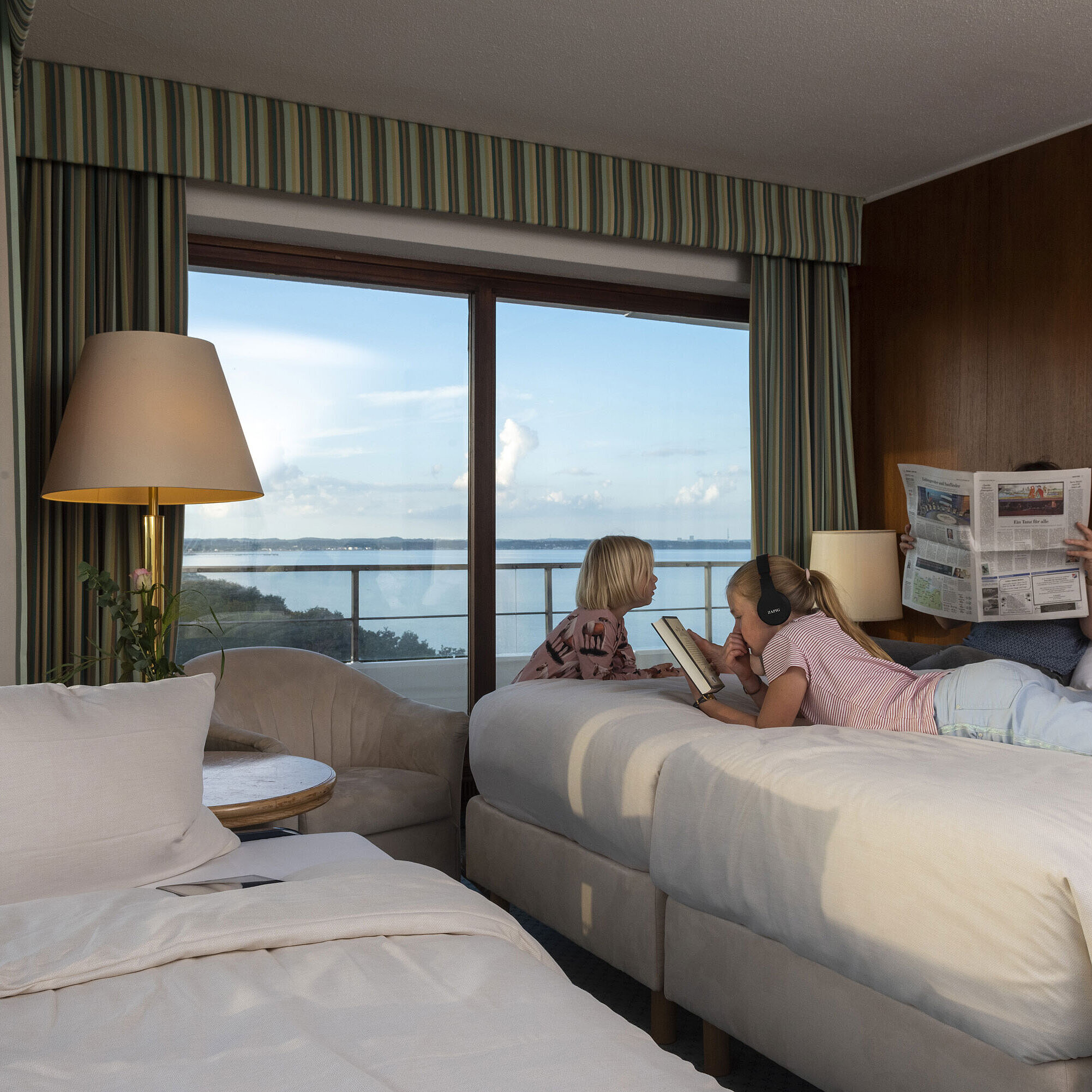 Familienzimmer Parkseite Familienzimmer im Maritim Hotel Timmendorfer Strand mit Kindern, die lesen, ein Tablet nutzen und den Meerblick genießen.