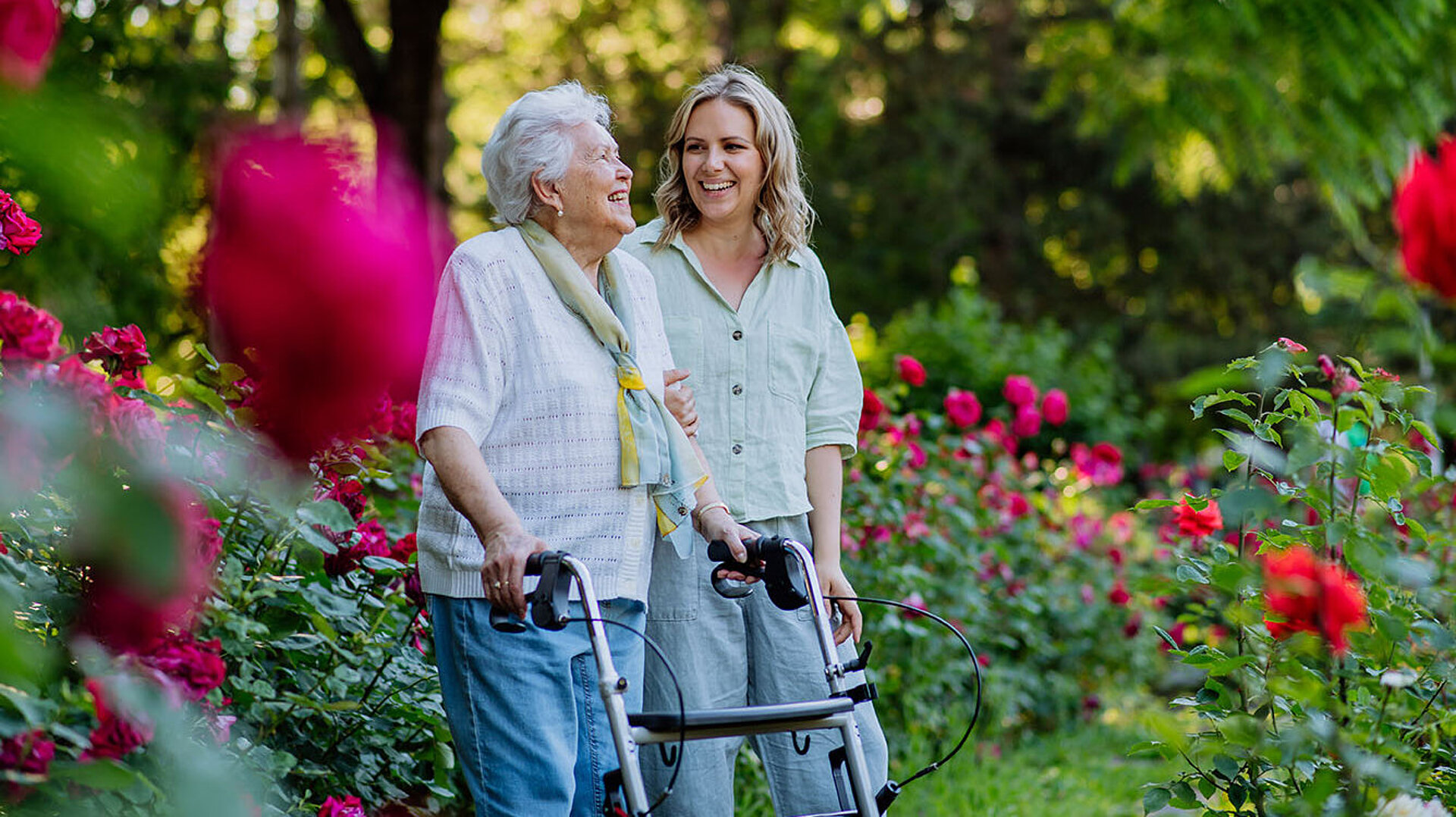 Zwei Frauen spazieren mit Rollator durch einen blühenden Rosengarten im Grünen.