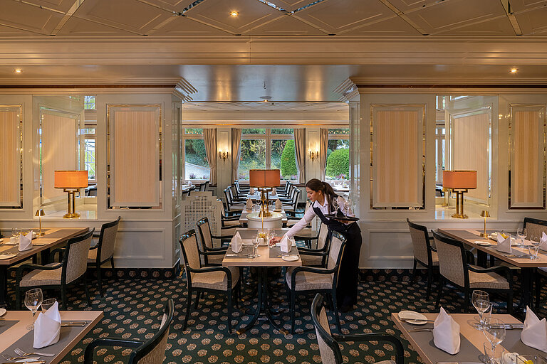 Restaurant Maritim Elegantes Restaurant mit eingedeckten Tischen, große Fenster und Servicekraft beim Vorbereiten des Gastraums.