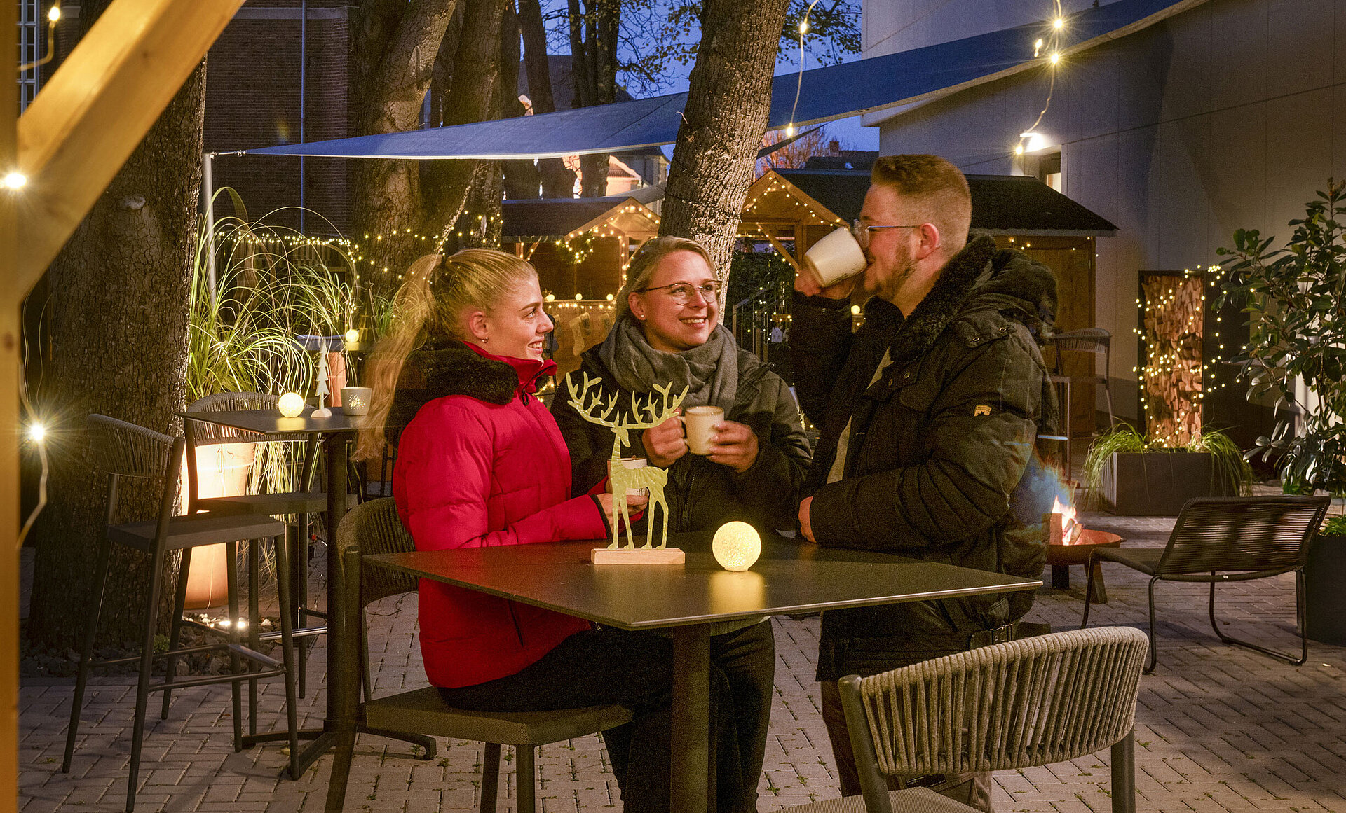 Maritim Weihnachtsdorf Drei Personen genießen heiße Getränke auf dem festlich geschmückten Weihnachtsmarkt im Maritim Darmstadt mit Lichterketten.