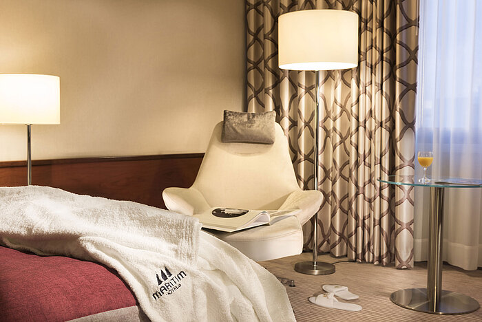 Detail eines Comfort Zimmers im Maritim Hotel Bonn mit Sessel, Tisch, Saftglas und Bademantel auf dem Bett.