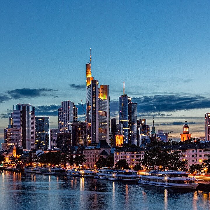 Blick über den Main auf die beleuchtete Skyline von Frankfurt am Main in der Abenddämmerung.