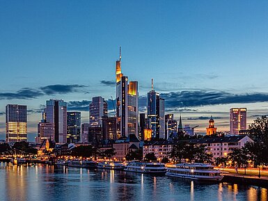 Blick über den Main auf die beleuchtete Skyline von Frankfurt am Main in der Abenddämmerung.