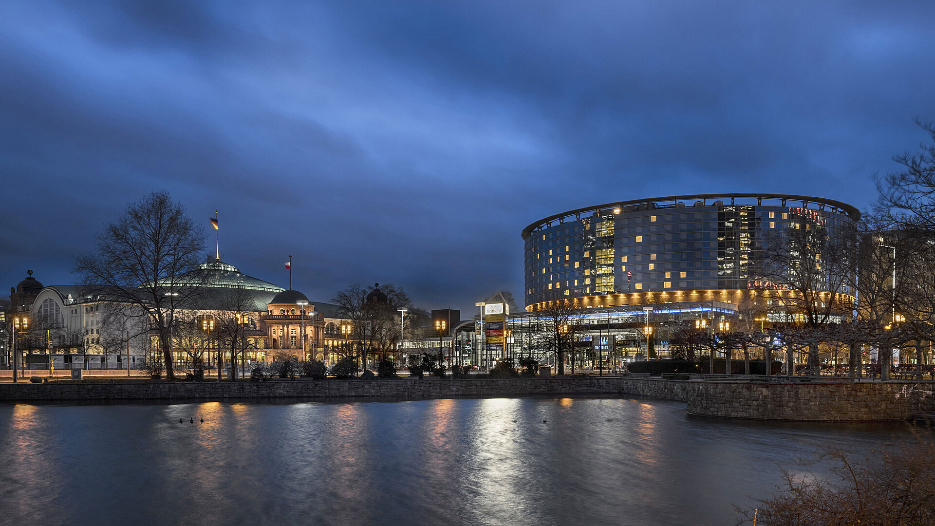 Maritim Hotel Frankfurt und Festhalle bei Nacht mit beleuchteter Glasfassade und Spiegelung im angrenzenden Teich.
