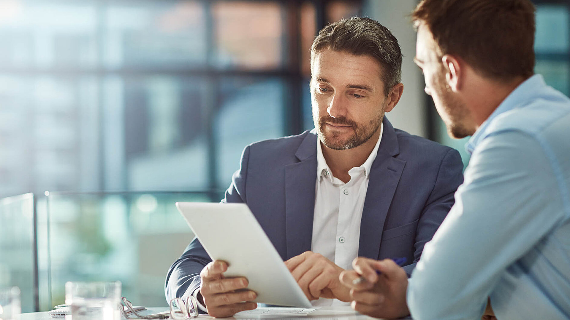 © Grady Rpeopleimages - Adobestock.com Zwei Geschäftsmänner im Büro besprechen Unterlagen auf einem Tablet.