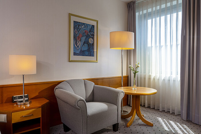Helles Hotelzimmer im Maritim Airport Hotel Hannover mit Sessel, Lampe, Fenster und Kunst.